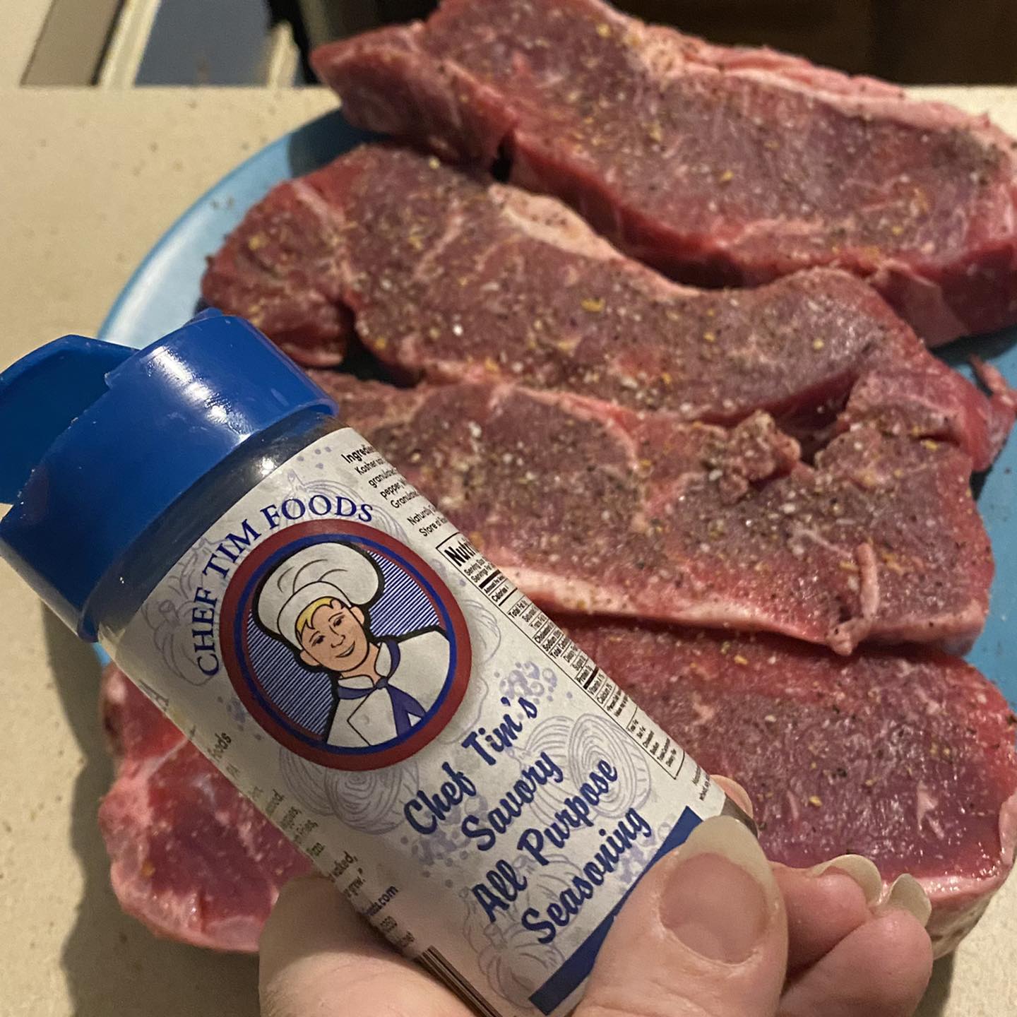 Hand holding a container of 'Chef Tim Foods' seasoning over raw meat on a blue plate.  Tim's Savory All Purpose Seasoning. A delightful blend of spices for beef, pork, chicken, seafood, veal & lamb, including roasts, steaks, chops, loins, fillets, and burgers. Also great on roasted potatoes, fries, pizza, vegetables & soups.
"Anything that walked, swam flew or grew."
Gluten Free & Cholesterol Free & No MSG
Great as a Gift for anyone who loves to Cook, Grill or use in the Smoker. Made in USA